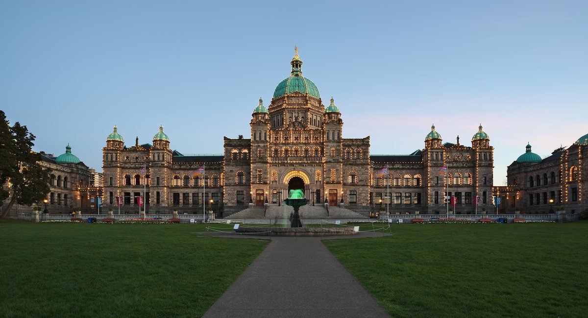 The domed stone Parliament Buildings in Victoria lit with strings of lights at dusk, a fountain and lawn in front.