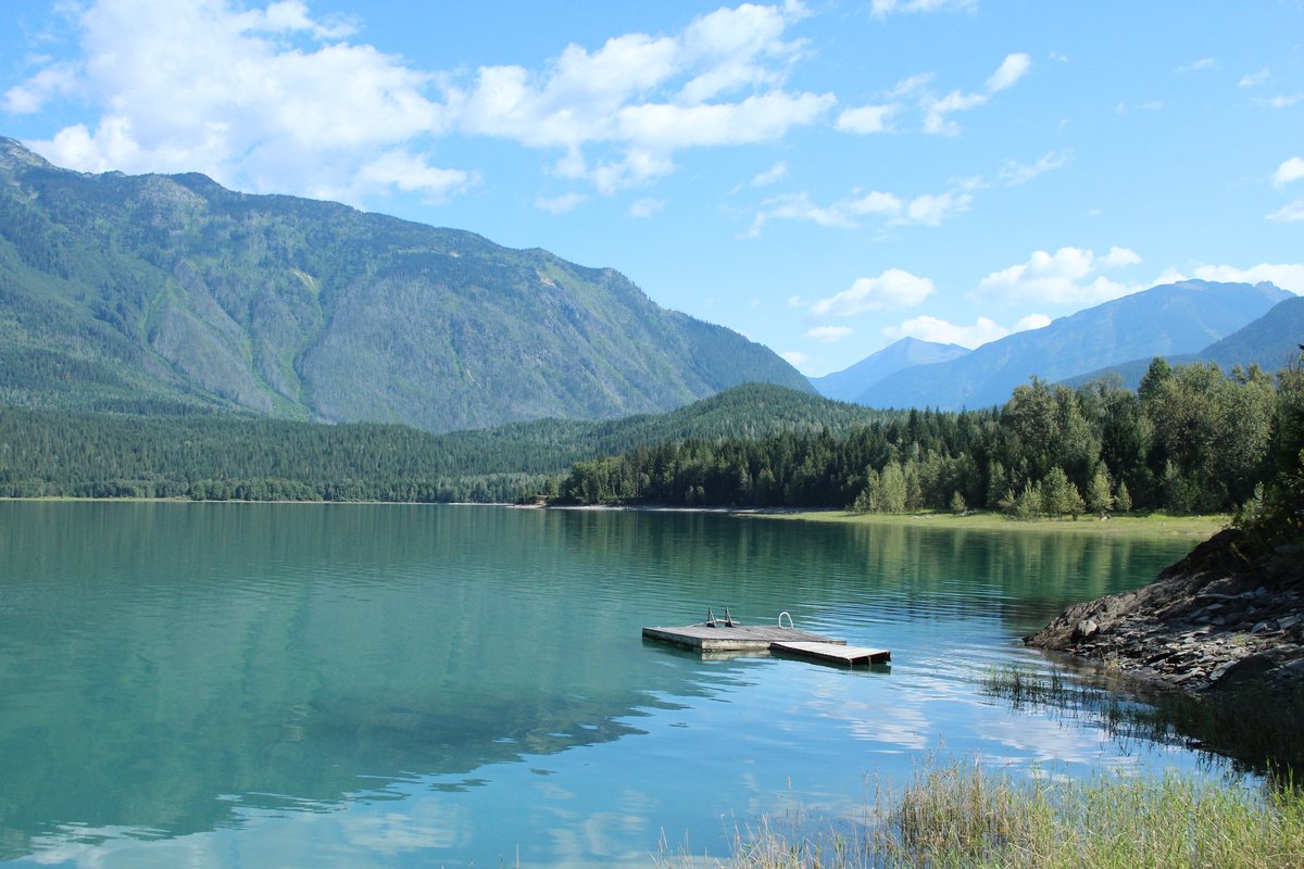 A swimming dock floats on the clear green water of Beaton Arm, with forested mountains and a valley opening beyond.
