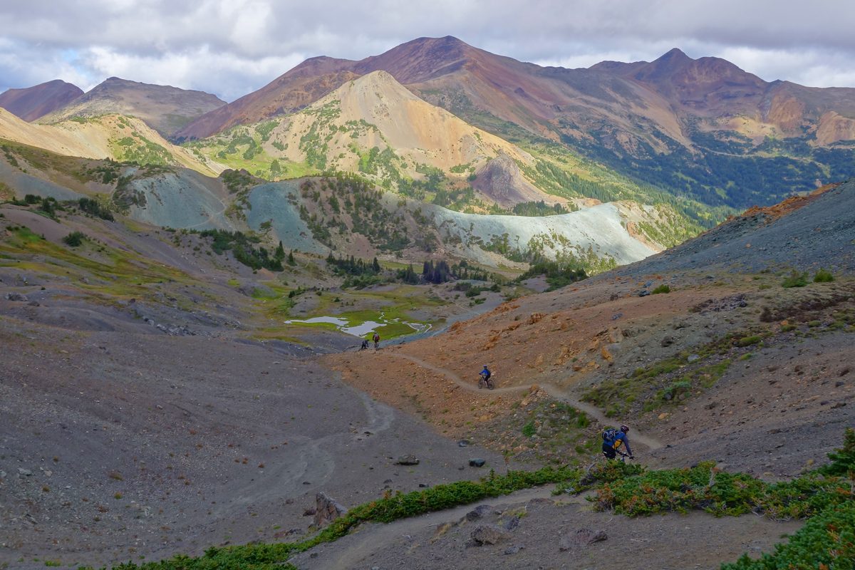 Mountain bikers riding a narrow trail across a bare, rust-coloured mountain valley in the Rainbow Range, with green meadows and peaks beyond.