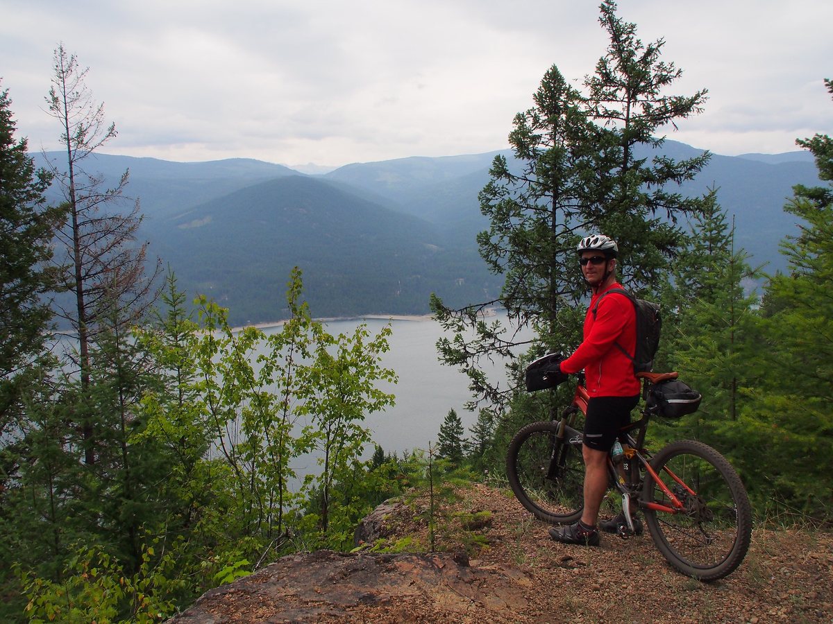 A cyclist in a red jacket stands with his bike on the old rail grade high above Lower Arrow Lake, with forested mountains beyond.
