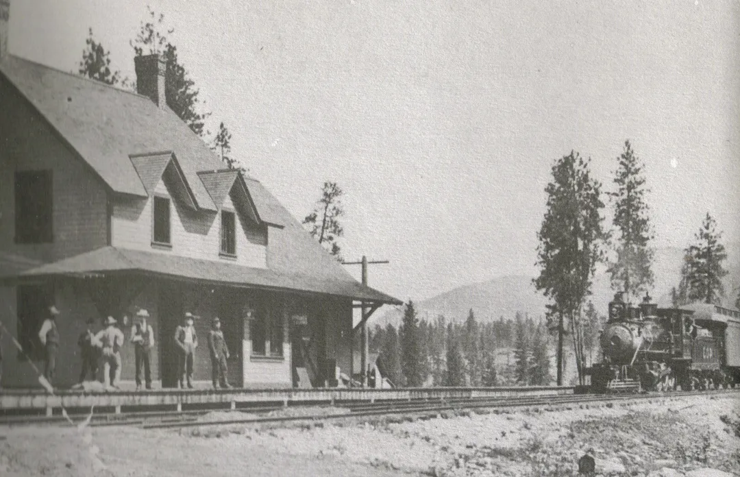 A black-and-white photograph from 1899 of the Columbia and Western Railway station at Cascade, near Christina Lake: a two-storey wooden station with dormer windows, a group of men in hats on the platform, and a steam locomotive standing at the right with pine-covered hills behind.