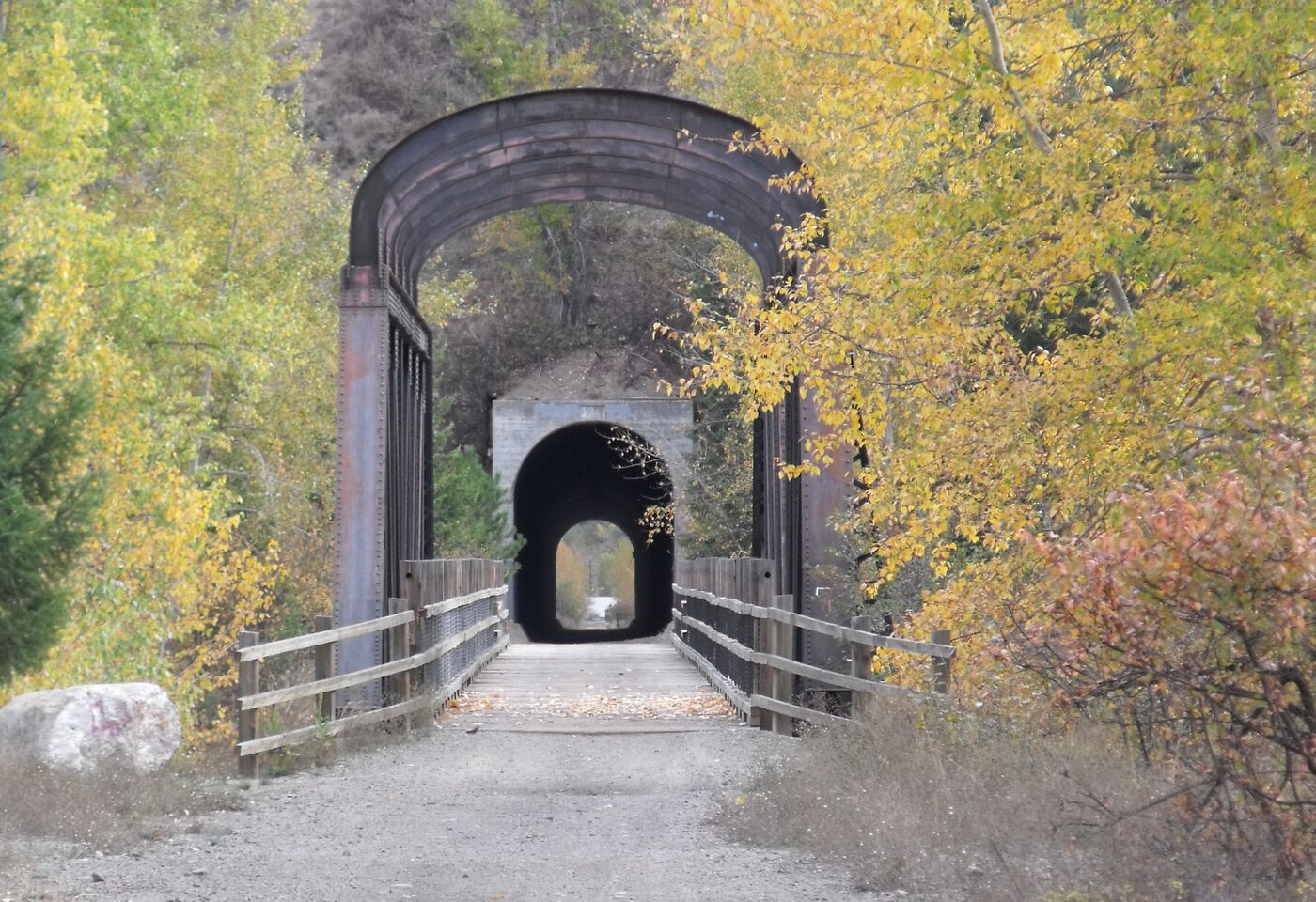 A gravel rail trail runs straight onto a steel through-truss bridge with an arched portal, and beyond it the dark mouth of a rock tunnel, framed by yellow autumn aspens and a dry hillside.