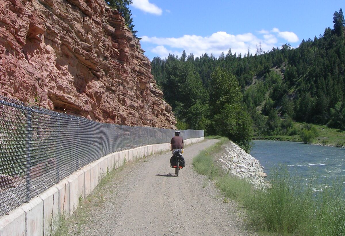 A loaded touring cyclist rides a gravel rail grade between a red rock cliff behind a chain-link catch fence and the Tulameen River.