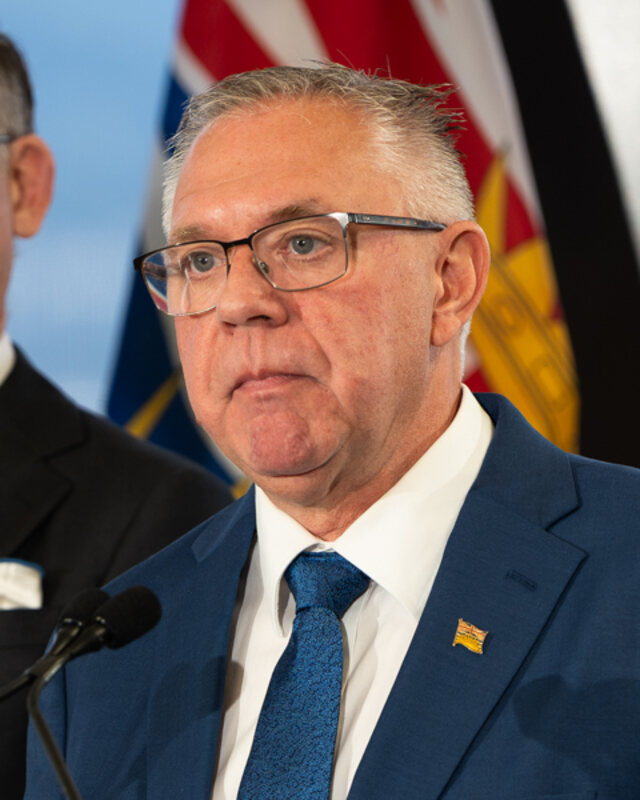 Lorne Doerkson at a lectern in a blue suit and tie, with the British Columbia flag behind him, at a campaign announcement on 28 September 2026.
