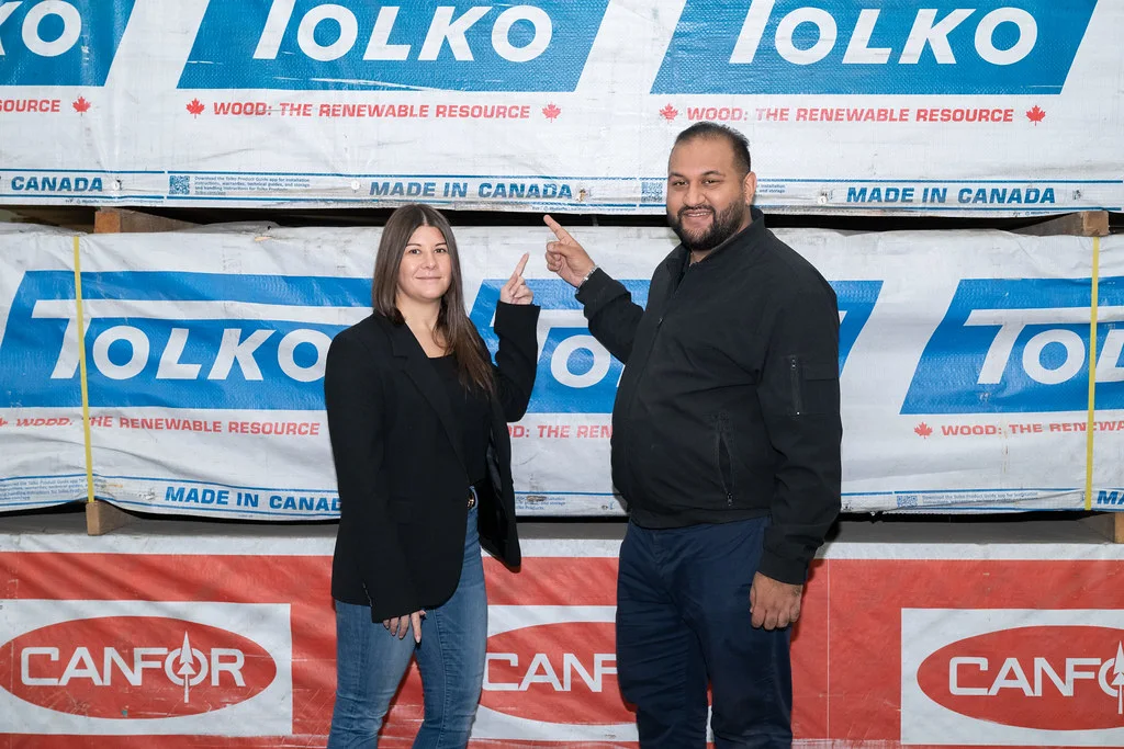 Ravi Parmar standing beside banded stacks of Tolko and Canfor lumber in a mill yard.