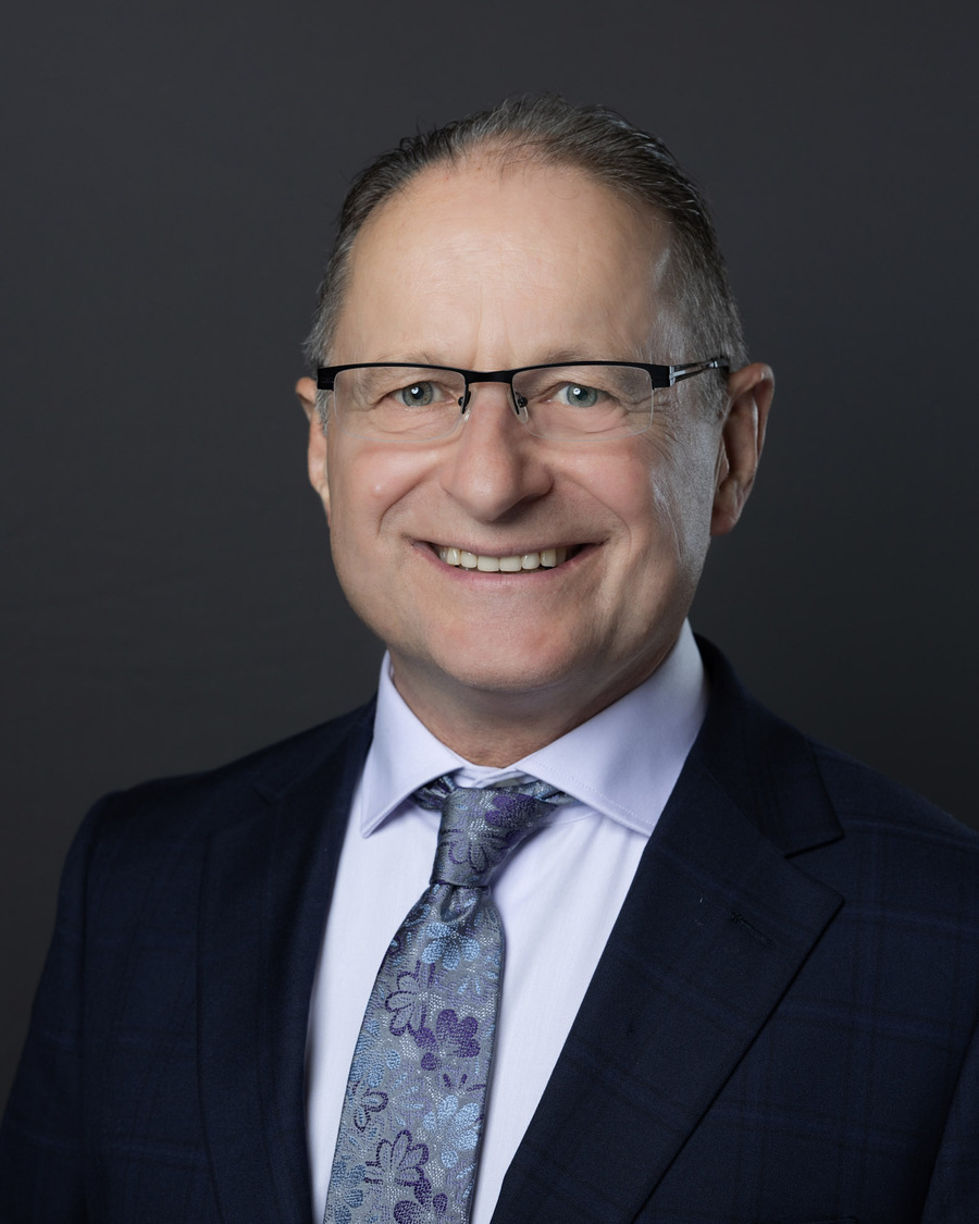 Official portrait of Steve Morissette: a studio headshot against a dark grey background, in a navy suit and glasses.