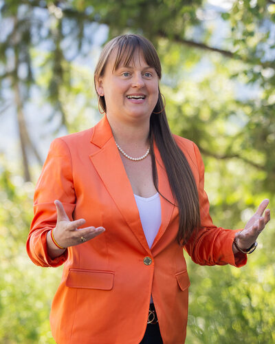 Brittny Anderson in an orange blazer, speaking outdoors in Nelson on 22 June 2026 with her hands open, trees behind her.