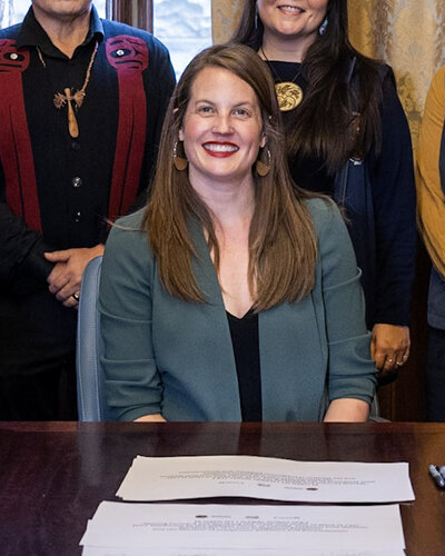 Christine Boyle in a green blazer, smiling, seated at a signing table in Victoria on 26 February 2025, when she was Minister of Indigenous Relations and Reconciliation.