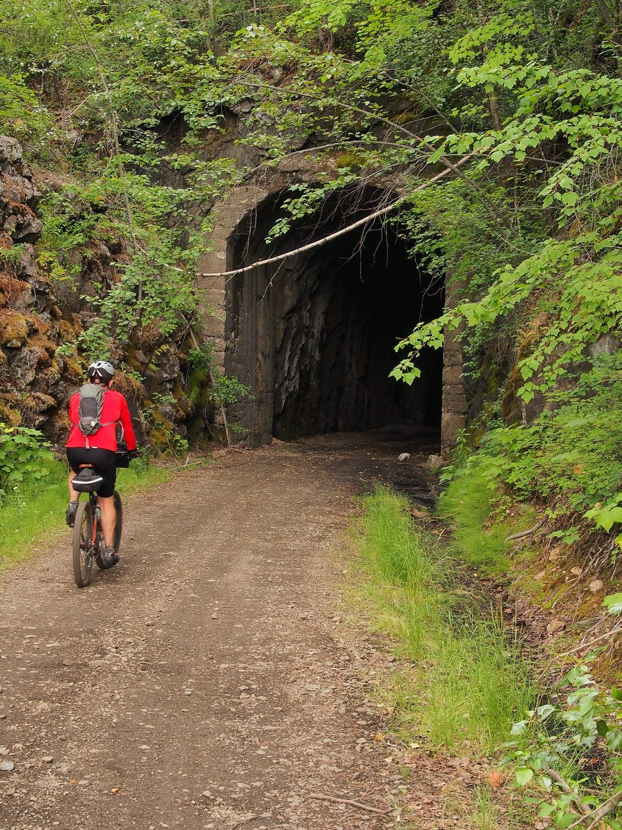 A cyclist in a red jacket rides along the gravel grade toward the dark mouth of the Bulldog Tunnel, cut into a mossy rock face.