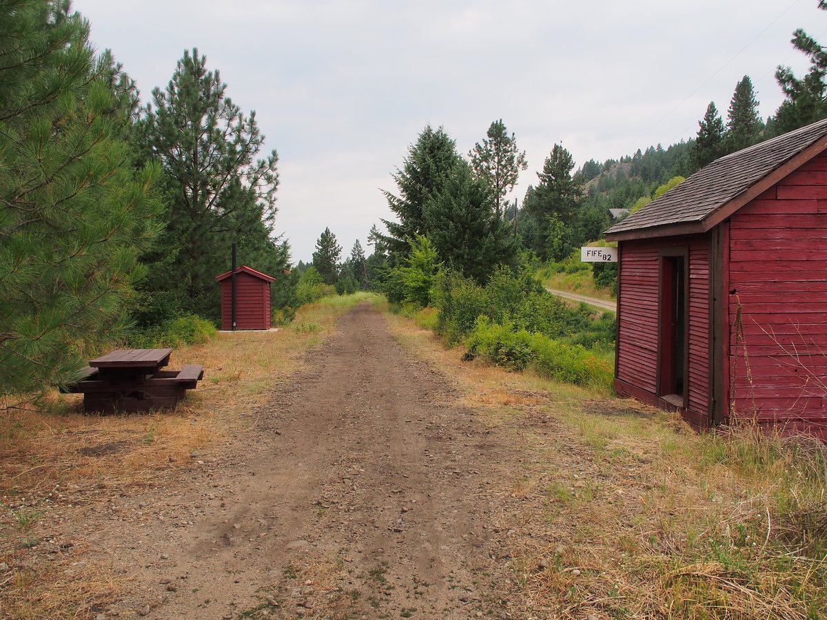 A gravel rail grade runs straight between conifers past a red trail shelter and a picnic table at the Fife trailhead near Christina Lake.