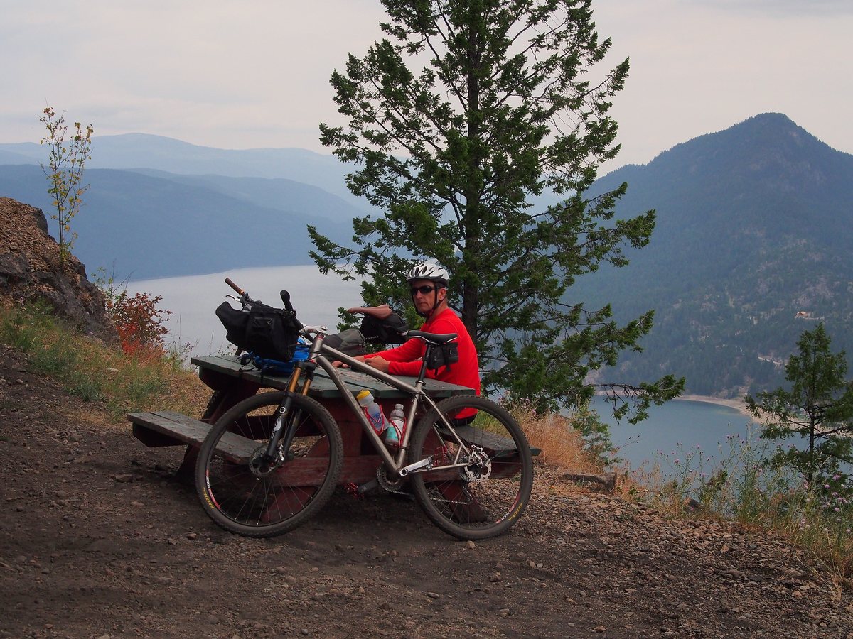 A cyclist sits at a picnic table beside his loaded bike on a viewpoint above Lower Arrow Lake.