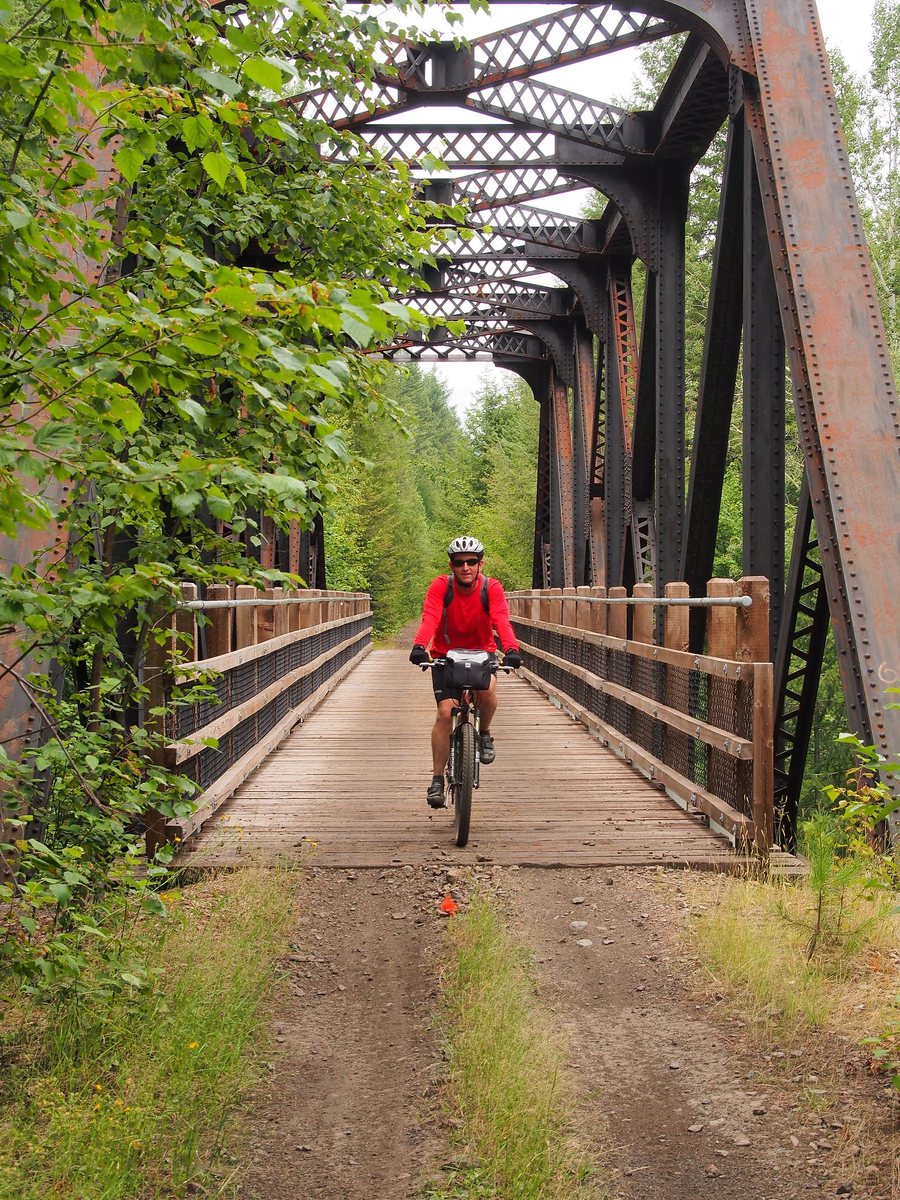 A cyclist in a red jacket rides across a steel truss bridge with a timber deck and wooden guardrails on the old rail grade.