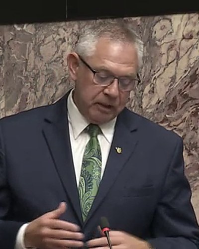 Lorne Doerkson, in a navy suit and green tie, speaking at his desk in the Legislative chamber as he introduced Bill M 247 on 26 May 2026.