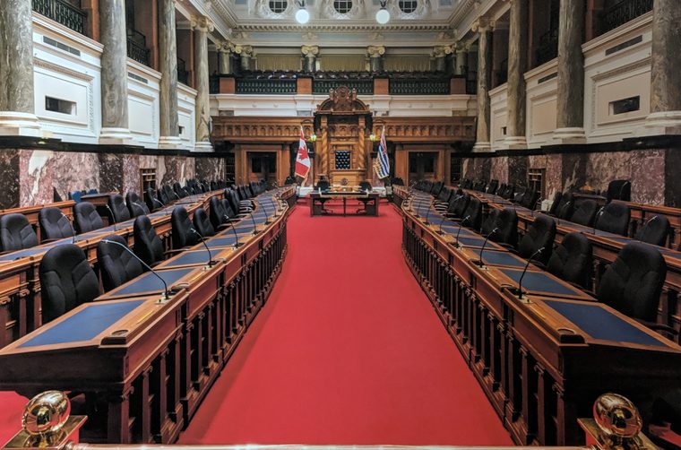 The empty legislative chamber of British Columbia: rows of wooden desks and black chairs on either side of a red carpet leading to the Speaker's canopied chair.