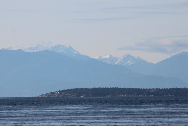 The low, wooded line of Discovery Island across open water, a small white lighthouse at its tip, with snow-capped mountains beyond.