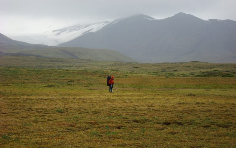 A lone hiker with a large pack crossing open alpine tundra, with cloud-capped mountains and a glacier in the distance.