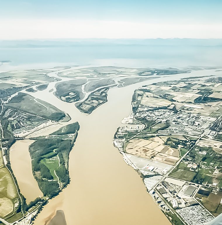 An aerial view of the wide brown south arm of the Fraser River, with Deas Island on the left and Richmond's farmland and industrial shore on the right.