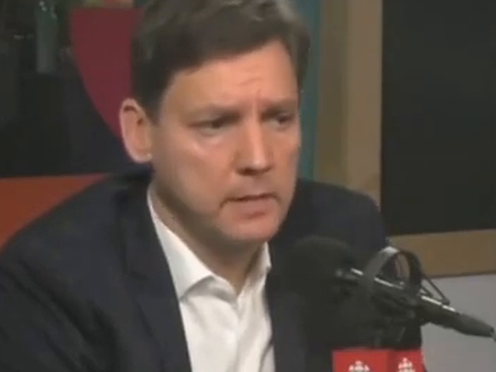 David Eby, in a dark suit and open white shirt, speaks into a microphone with CBC logos in a radio studio.