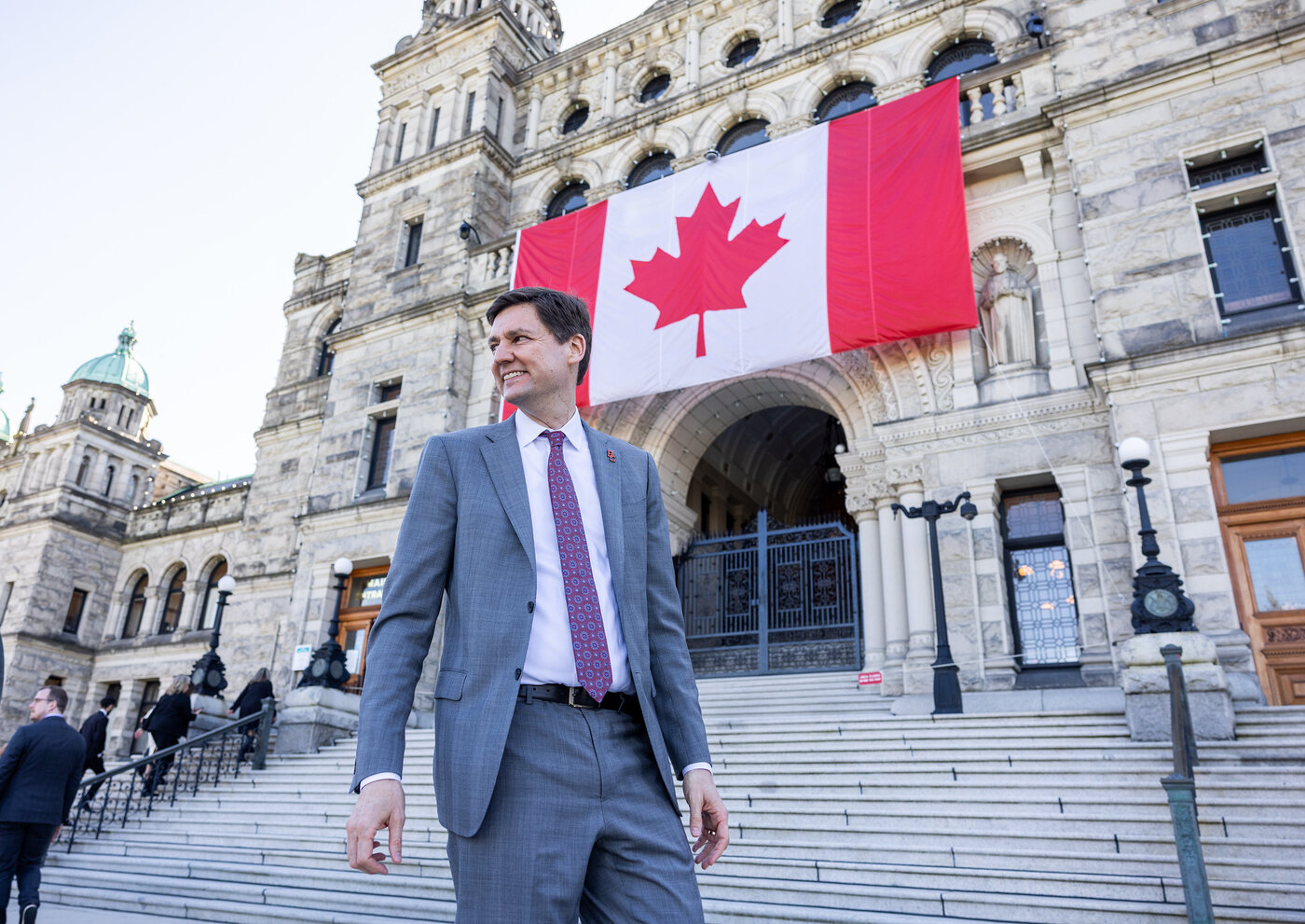 David Eby, smiling, in a grey suit and patterned tie, walking down the steps of the B.C. Legislature with a large Canadian flag hung over the entrance behind him.