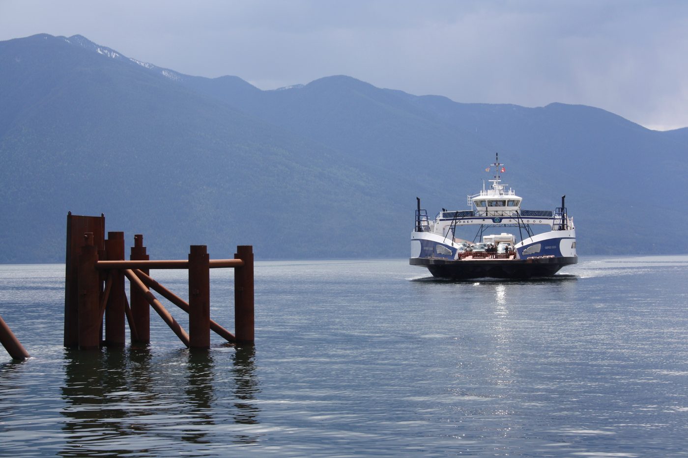The Kootenay Lake ferry crossing calm water toward a forested shore, with mountains rising behind.