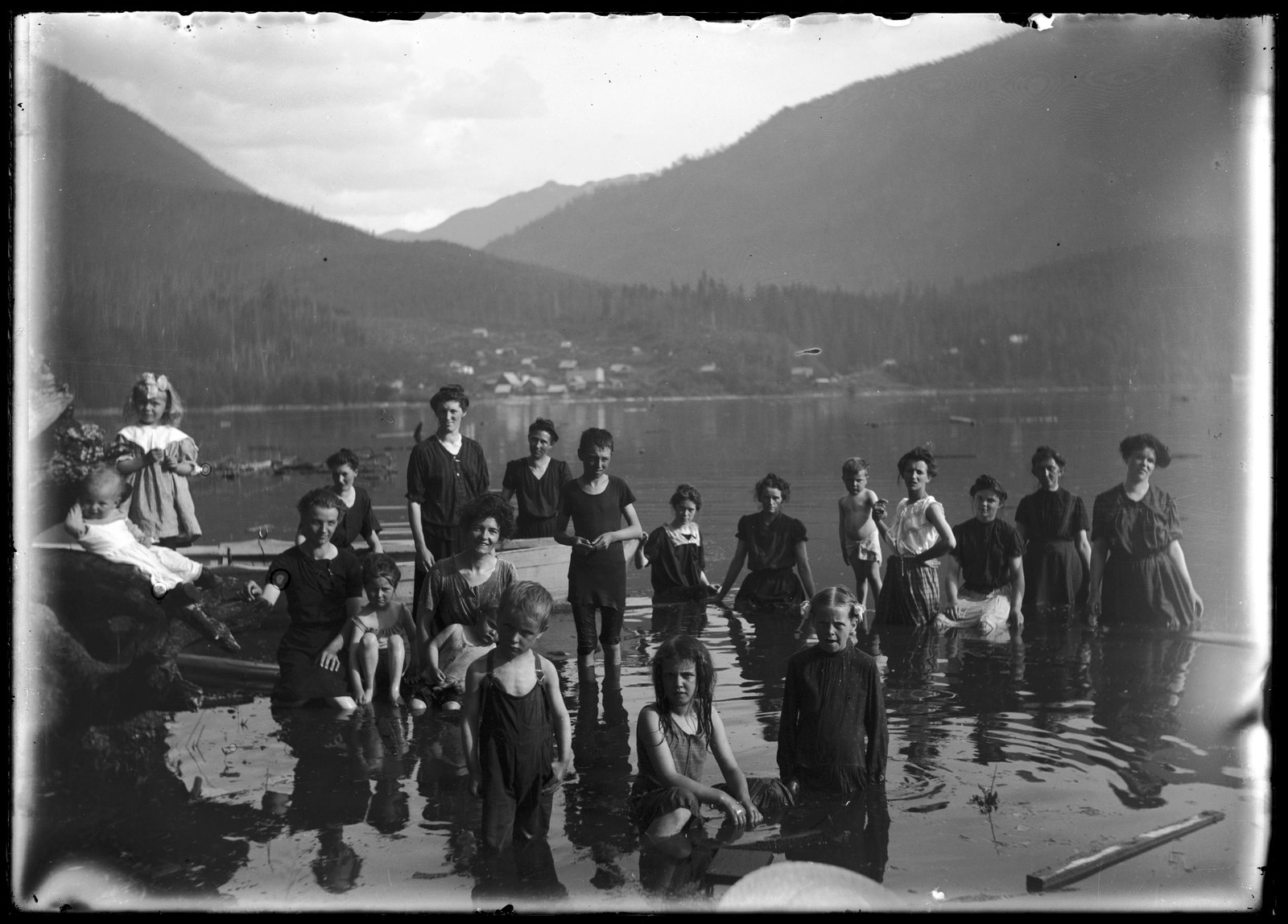 A black and white photograph of women and children standing waist-deep in a lake, with a small town and forested mountains across the water behind them.