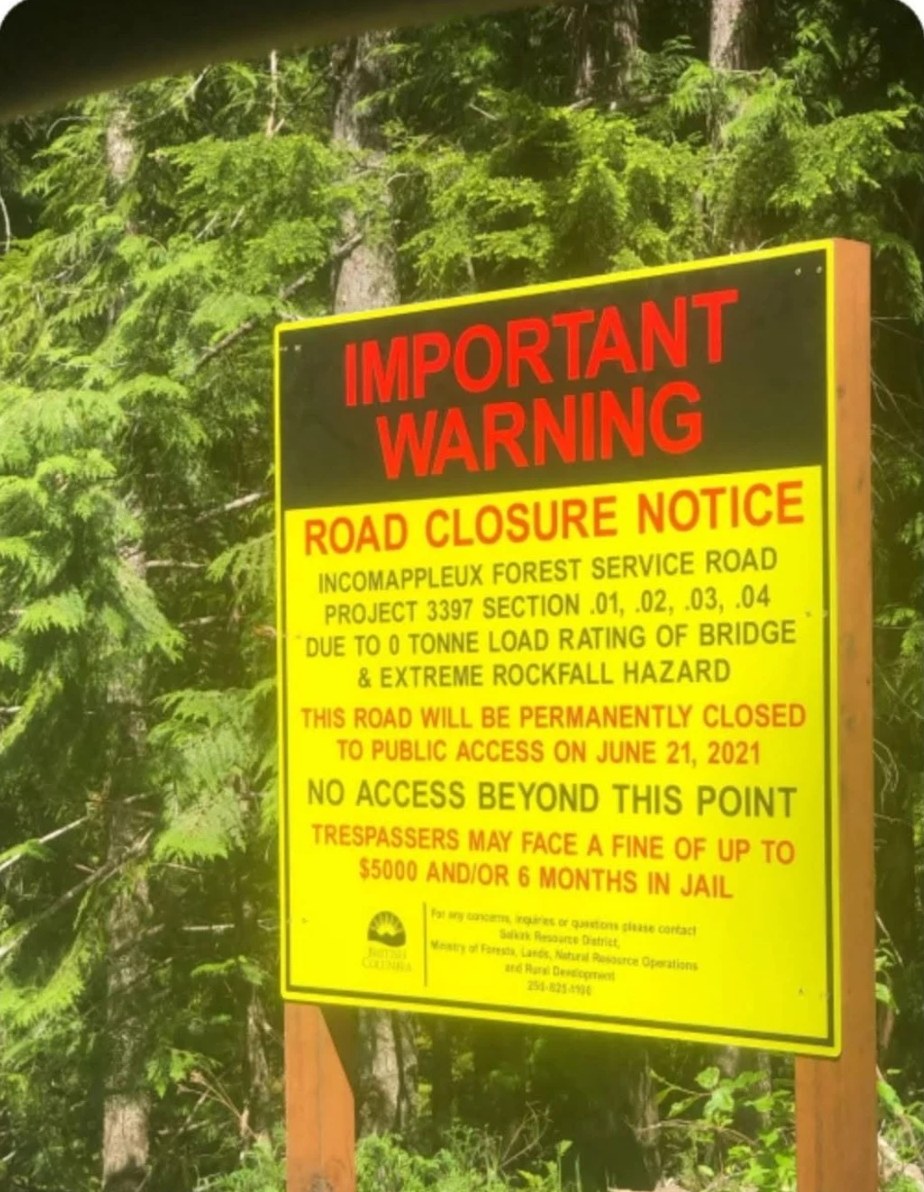 A yellow Province of British Columbia sign on two wooden posts in a green forest, headed Important Warning, Road Closure Notice, Incomappleux Forest Service Road, saying the road will be permanently closed to public access on June 21, 2021, and that trespassers may face a fine of up to $5000 and/or 6 months in jail.