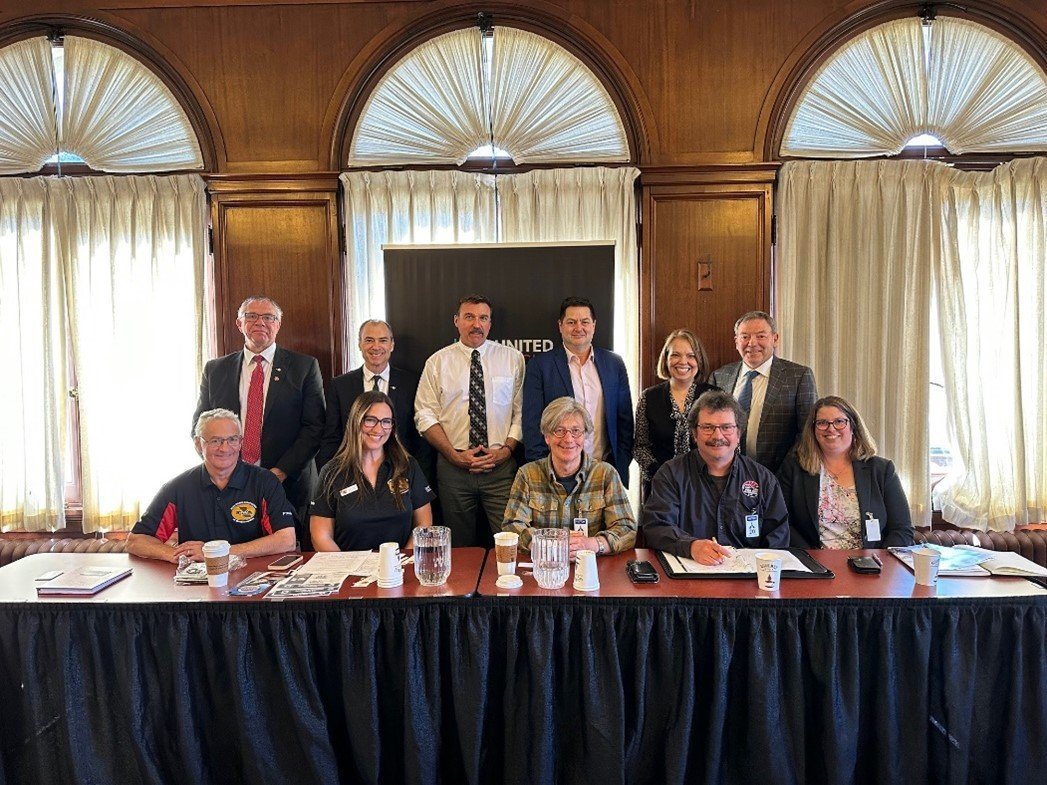Six BC United MLAs standing behind a table where representatives of four motorized recreation associations are seated, in a wood-panelled room in Victoria.