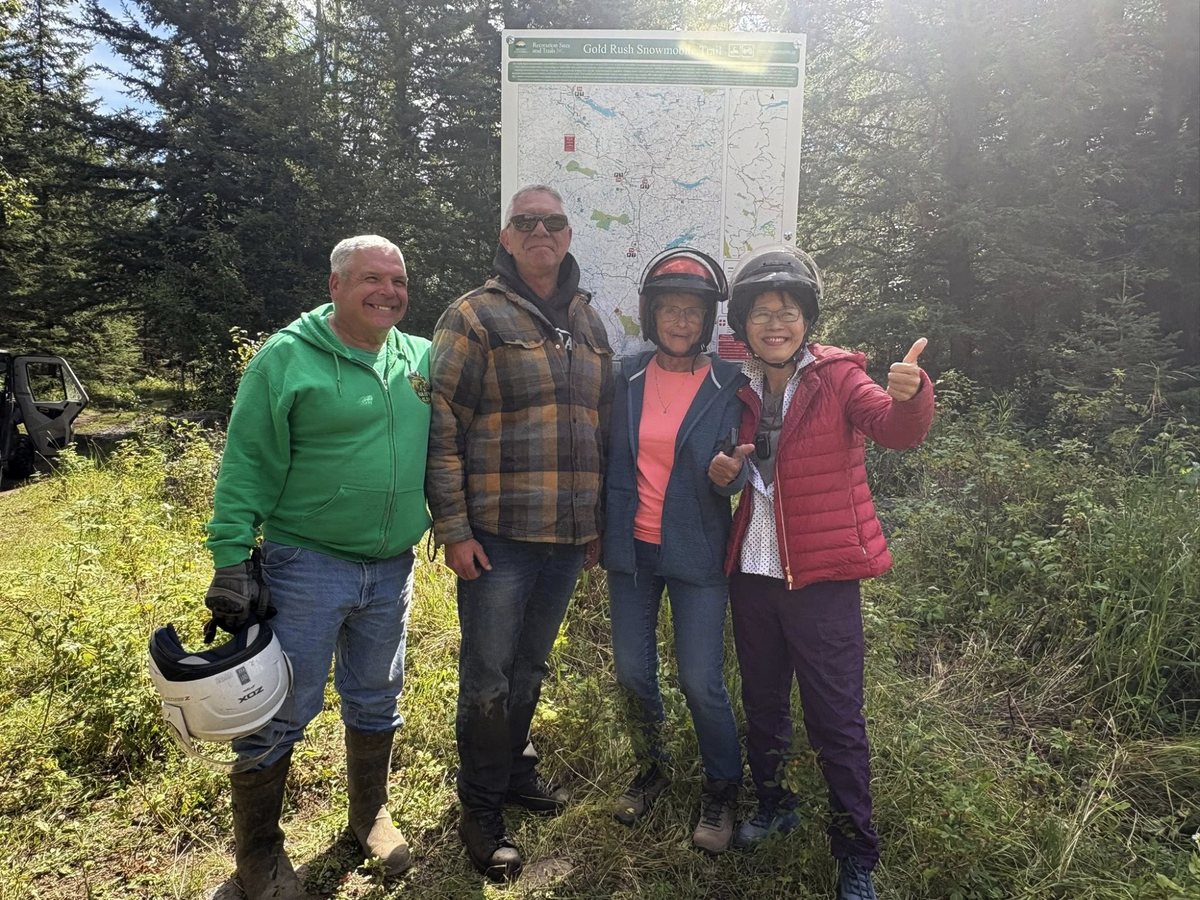 Four people stand in front of a trail map in the woods: Mark Redl in a green jacket, Lorne Doerkson in a plaid jacket, and two women in helmets, one giving a thumbs-up.