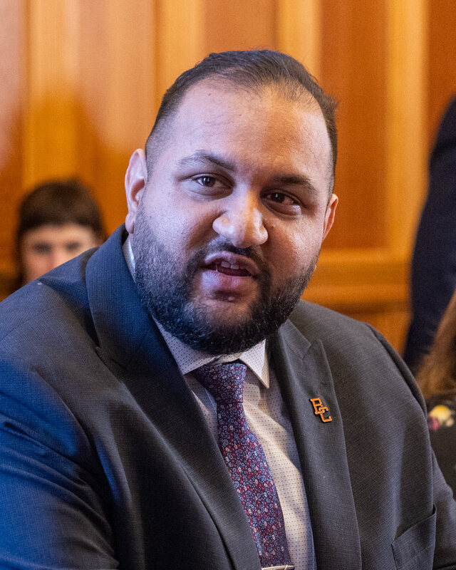 Ravi Parmar, in a dark suit and patterned tie, speaking at a table in the Legislature.
