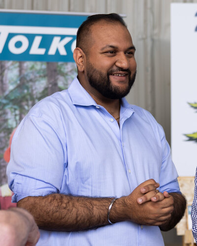 Ravi Parmar in a light blue shirt, smiling, with his hands clasped, in front of a Tolko banner at a mill in Kelowna on 11 June 2026.