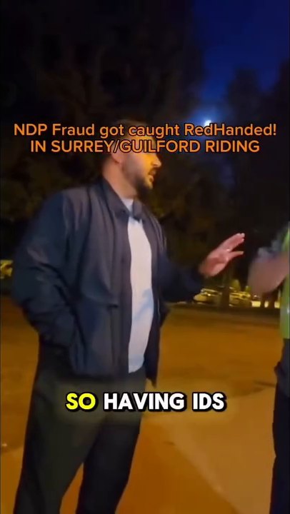 The man in the navy jacket raises a hand as he explains, in a dark parking lot with parked cars behind.