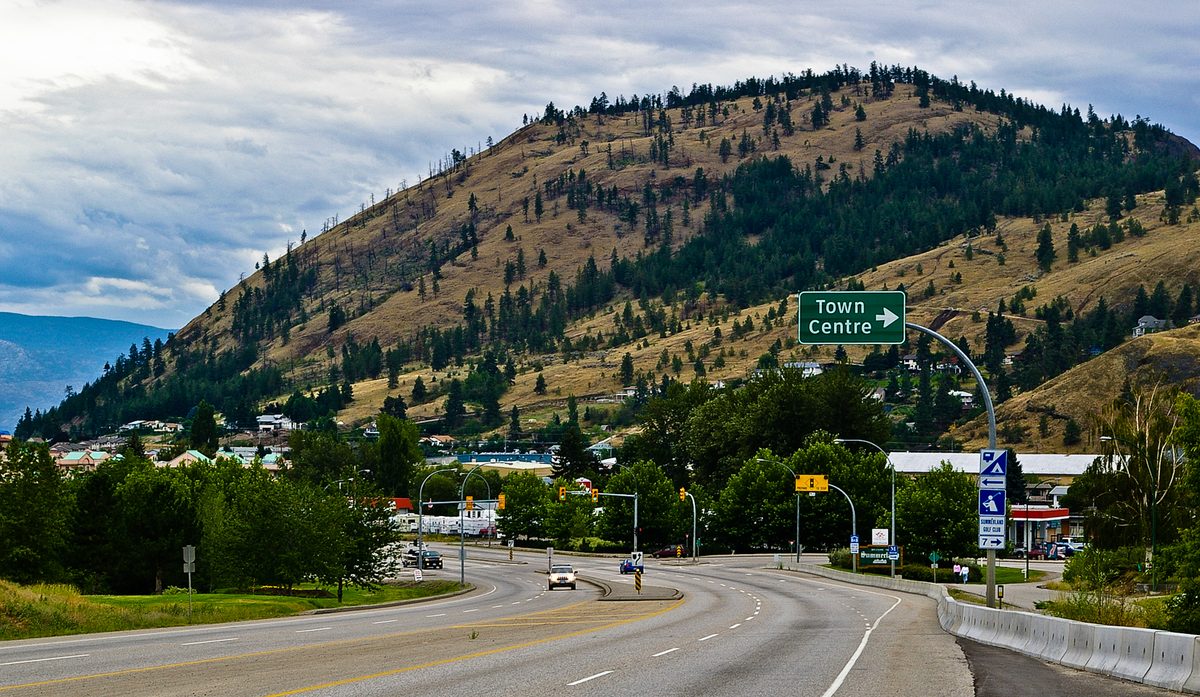 Highway 97 curving into Summerland under a dry, pine-dotted hill, with a green Town Centre sign over the road.
