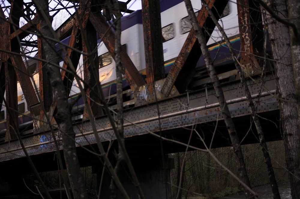 A West Coast Express train crossing a rusted steel railway bridge through bare trees.
