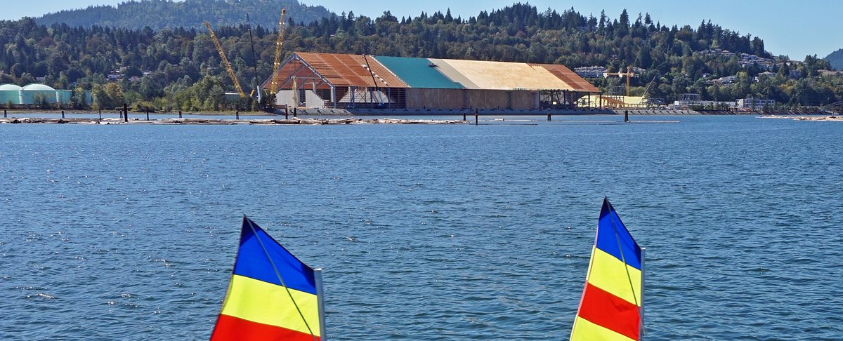 The head of Burrard Inlet at Port Moody: a mill on the far shore below forested hills, and the tips of two small sails in the foreground.