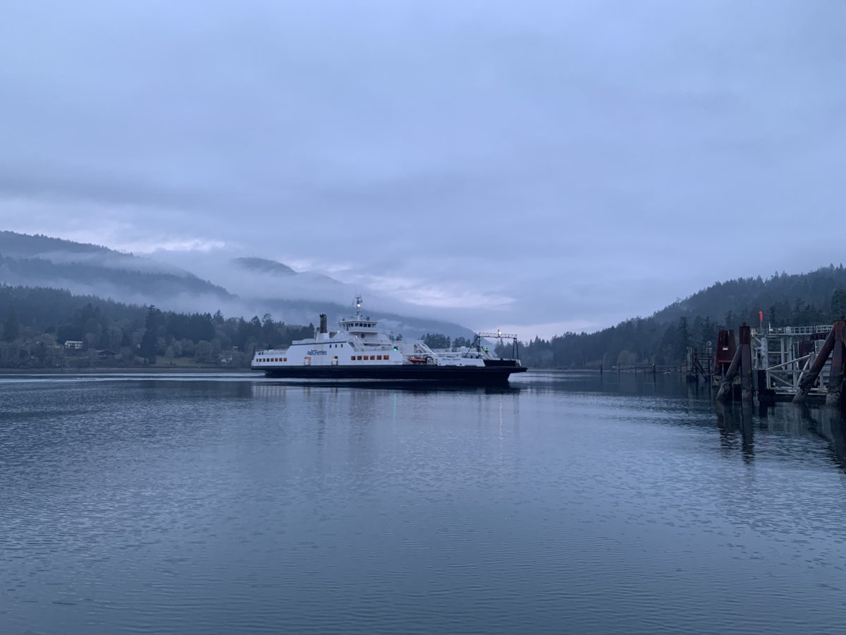 A white B.C. Ferries vessel crossing calm grey water toward Fulford Harbour, under low cloud on forested hills.