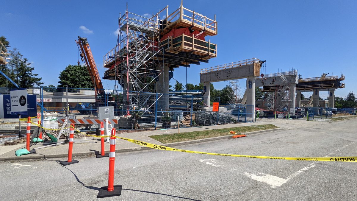 The Fleetwood SkyTrain station under construction beside Fraser Highway: concrete columns, scaffolding, a crane and caution tape.
