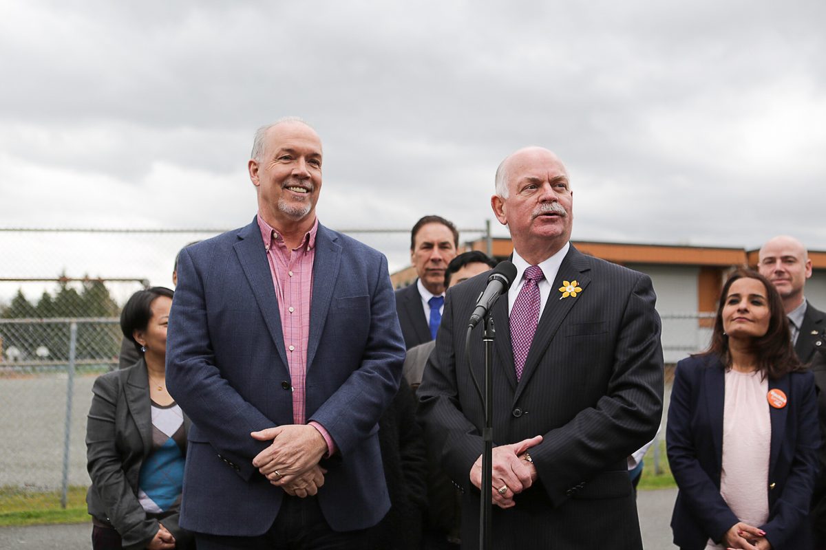 Garry Begg speaks at a microphone beside John Horgan in a Surrey schoolyard in April 2017, with a portable classroom building behind them.