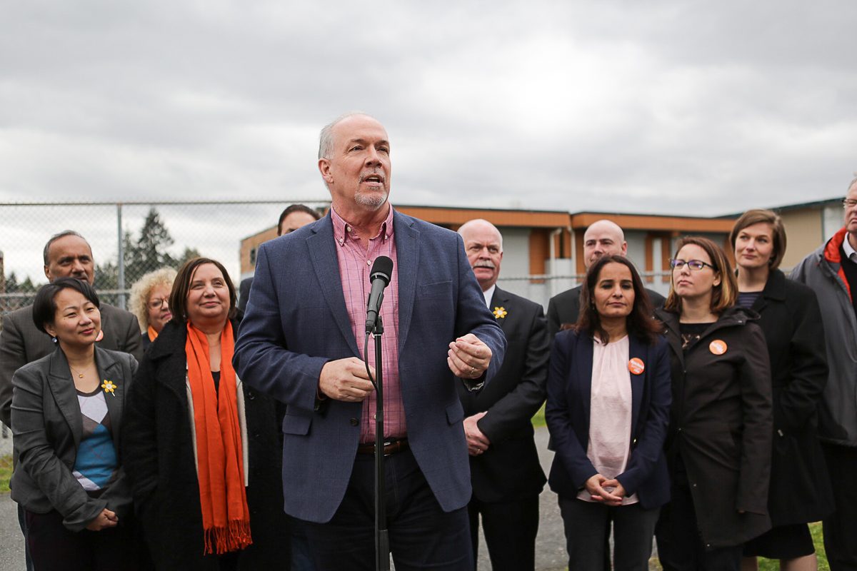 John Horgan speaks at a microphone in a Surrey schoolyard, with NDP candidates behind him and a portable classroom building in the background.