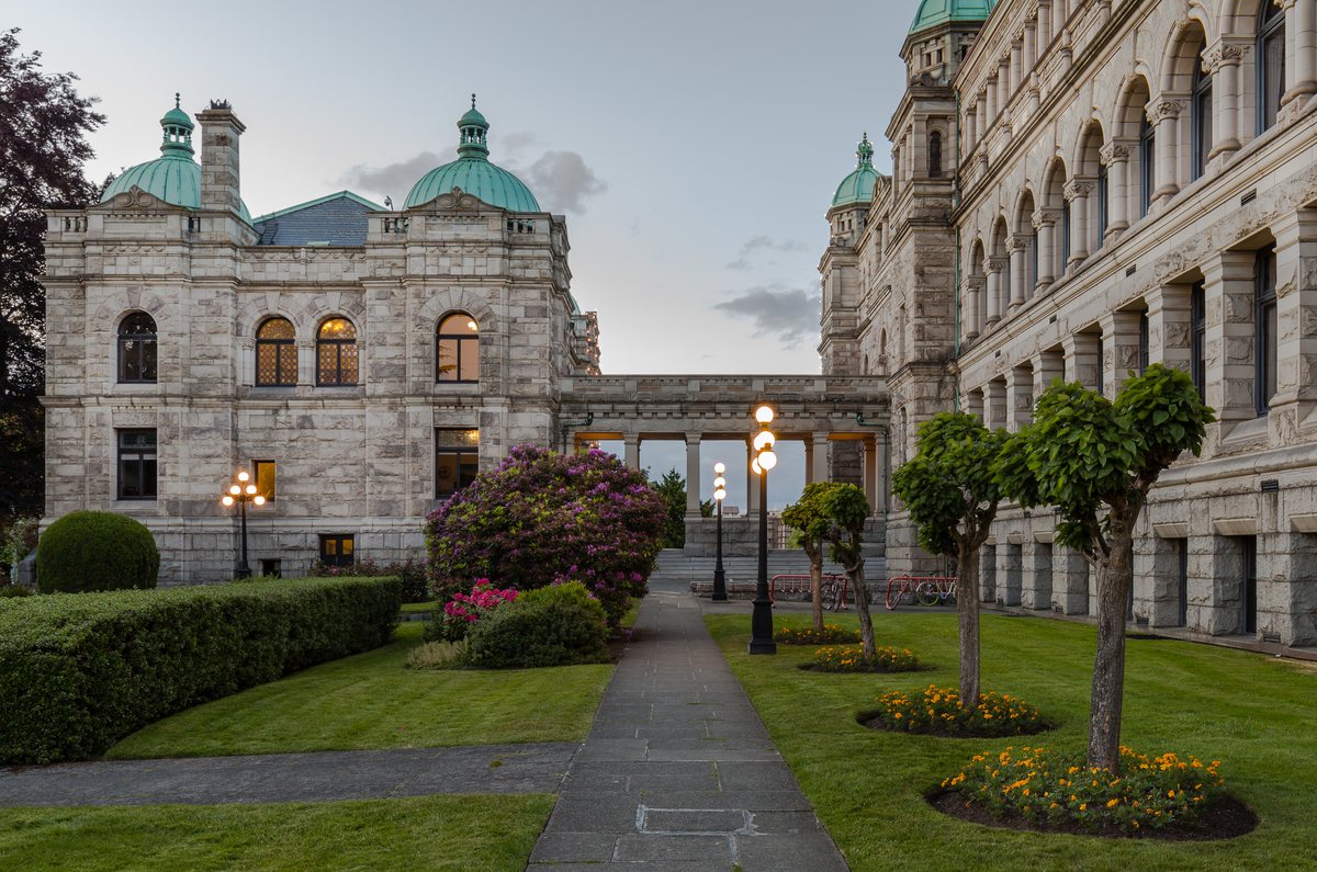 The lit stone portico and steps of the British Columbia Parliament Buildings at dusk, with flower beds in front.