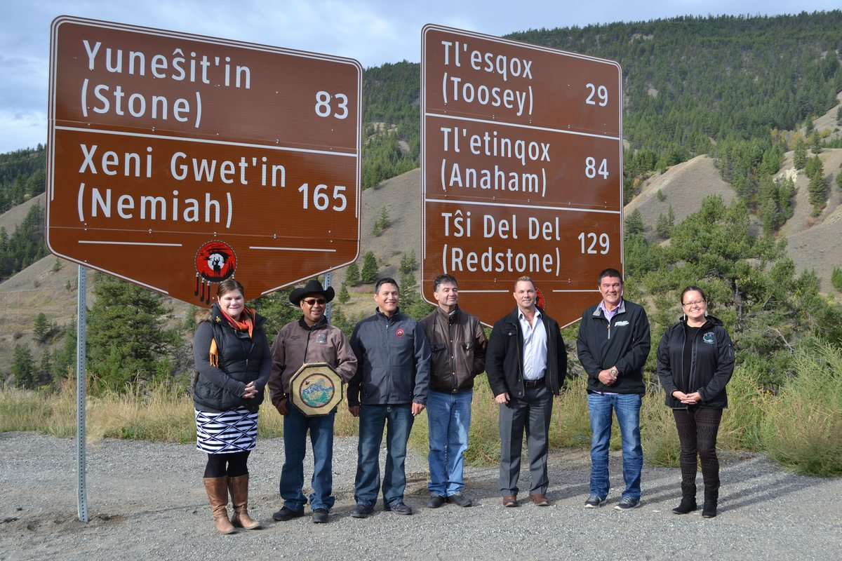 Seven people stand beneath two large brown highway signs listing Tsilhqot'in communities and distances, including Xeni Gwet'in (Nemiah) 165, on a gravel roadside with dry hills behind.